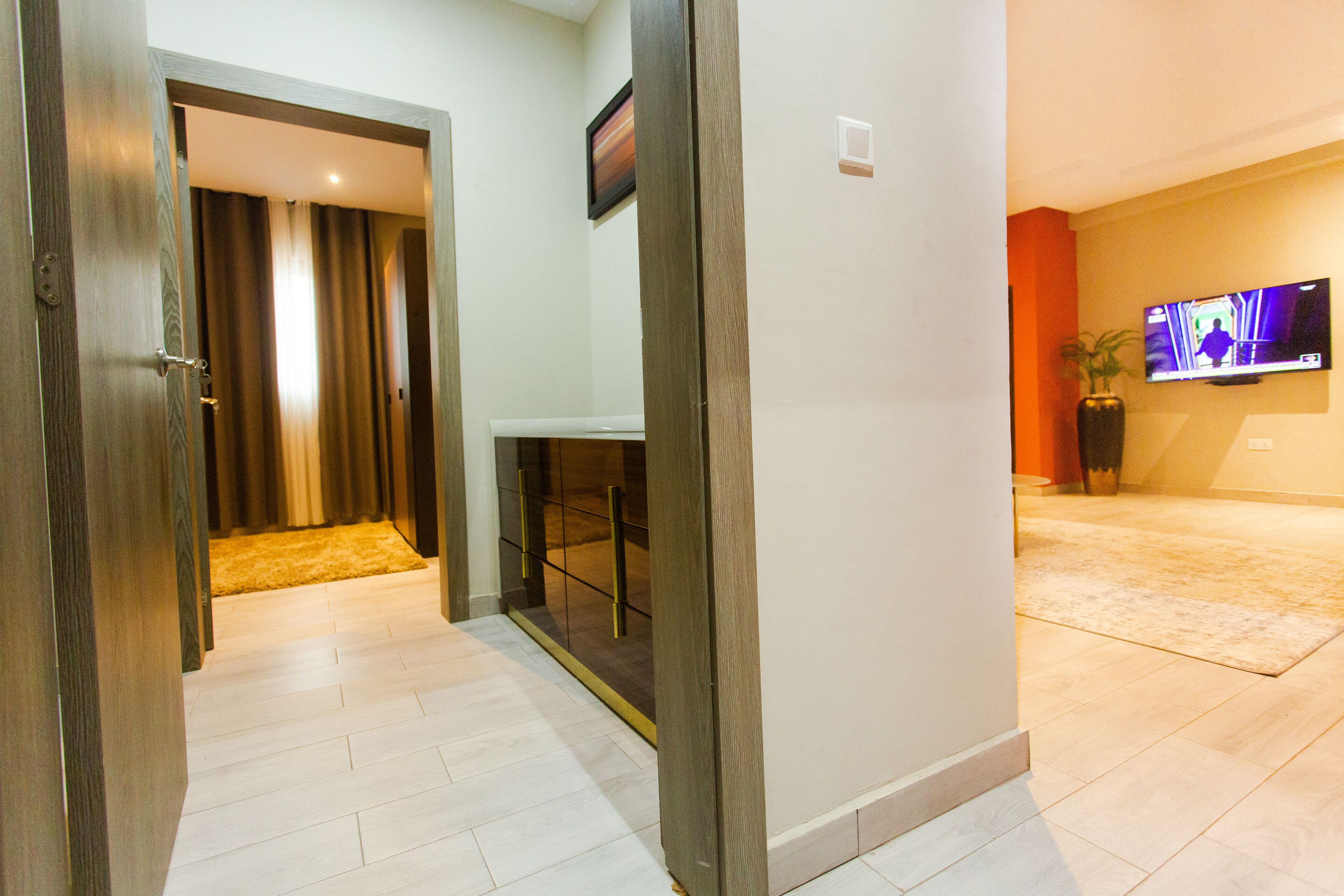 A modern hotel room with a wooden door, a small table, a TV mounted on the wall, and a potted plant.