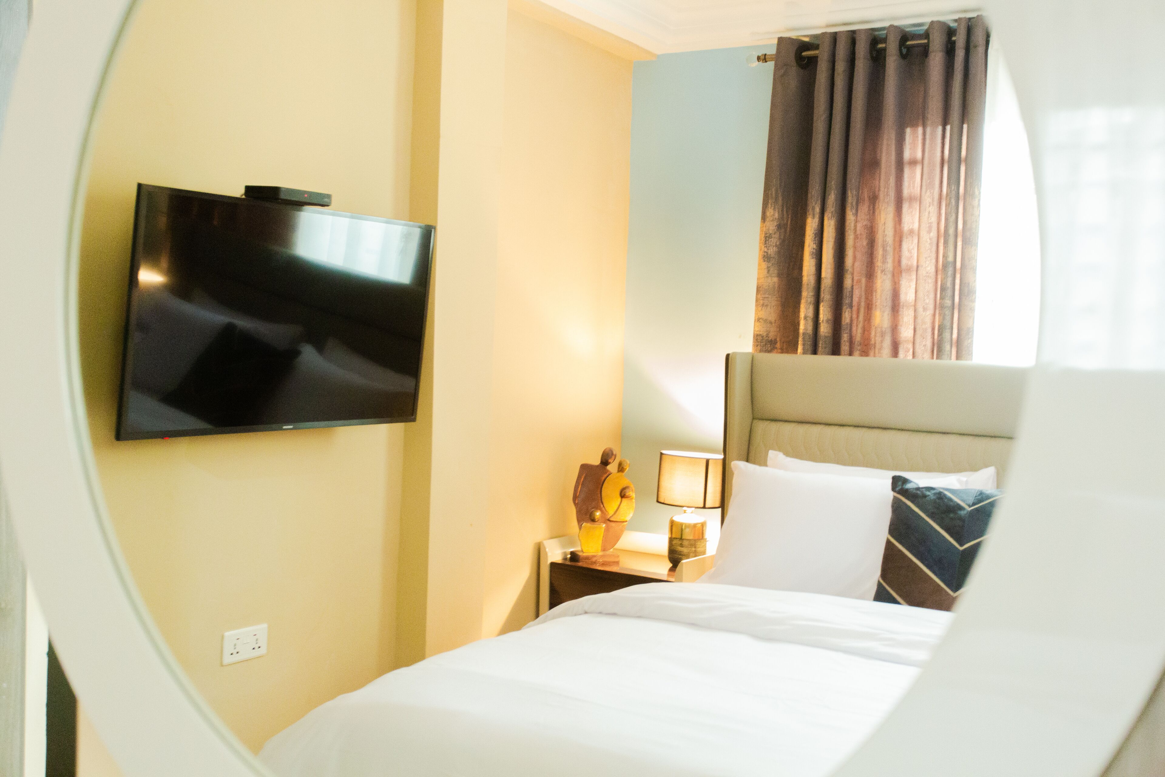 A hotel room with a bed, a flat-screen TV mounted on the wall, a nightstand with a lamp, and a window with curtains.