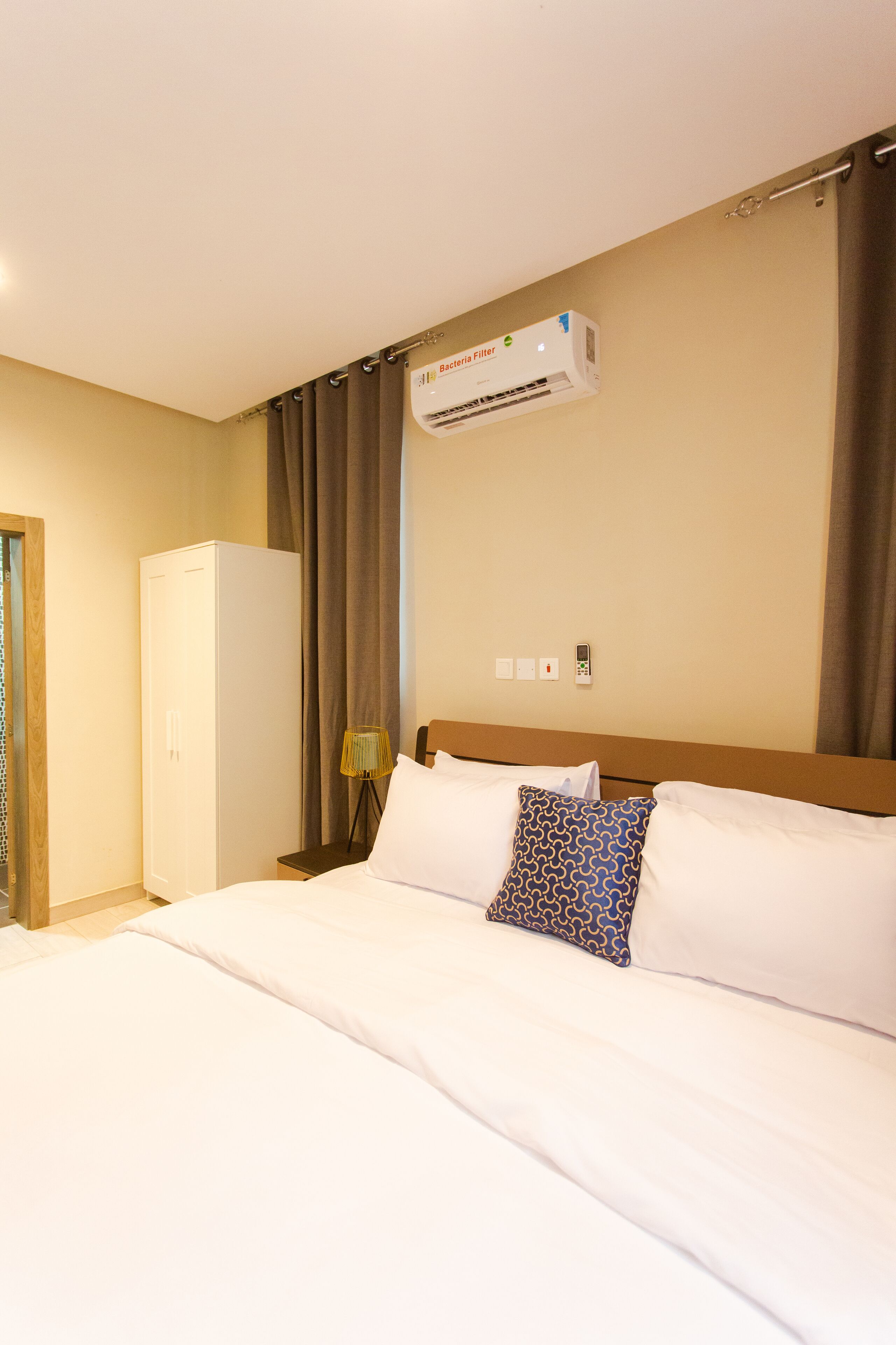 A hotel room with a bed, a wardrobe, an air conditioning unit, a lamp, and a wall-mounted phone.