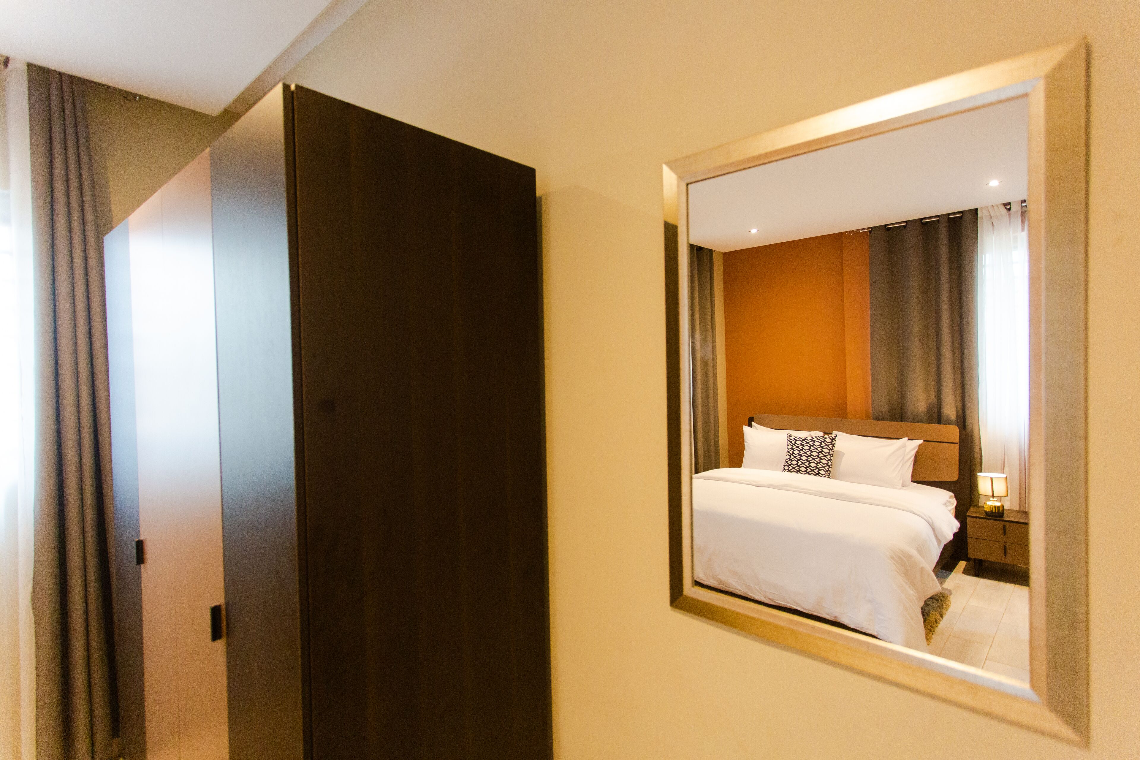 Habitación de hotel con un espejo grande que refleja una cama bien hecha, una mesita de noche de madera con una lámpara y un armario de color oscuro.
