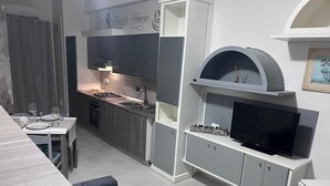 A compact kitchen with a gas stove, sink, and a built-in oven. There is a dining area with a table set for two and a sofa. A television is mounted on the wall above a cabinet. - Nina's house 3 300 meters from the sea (Santa Caterina Dello Ionio Marina)