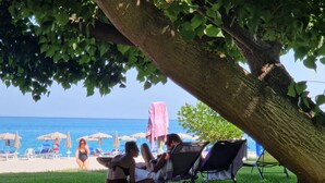 A beach scene with a tree providing shade, a grassy area, and beach chairs and umbrellas in the background. - Nina's house 3 300 meters from the sea (Santa Caterina Dello Ionio Marina)