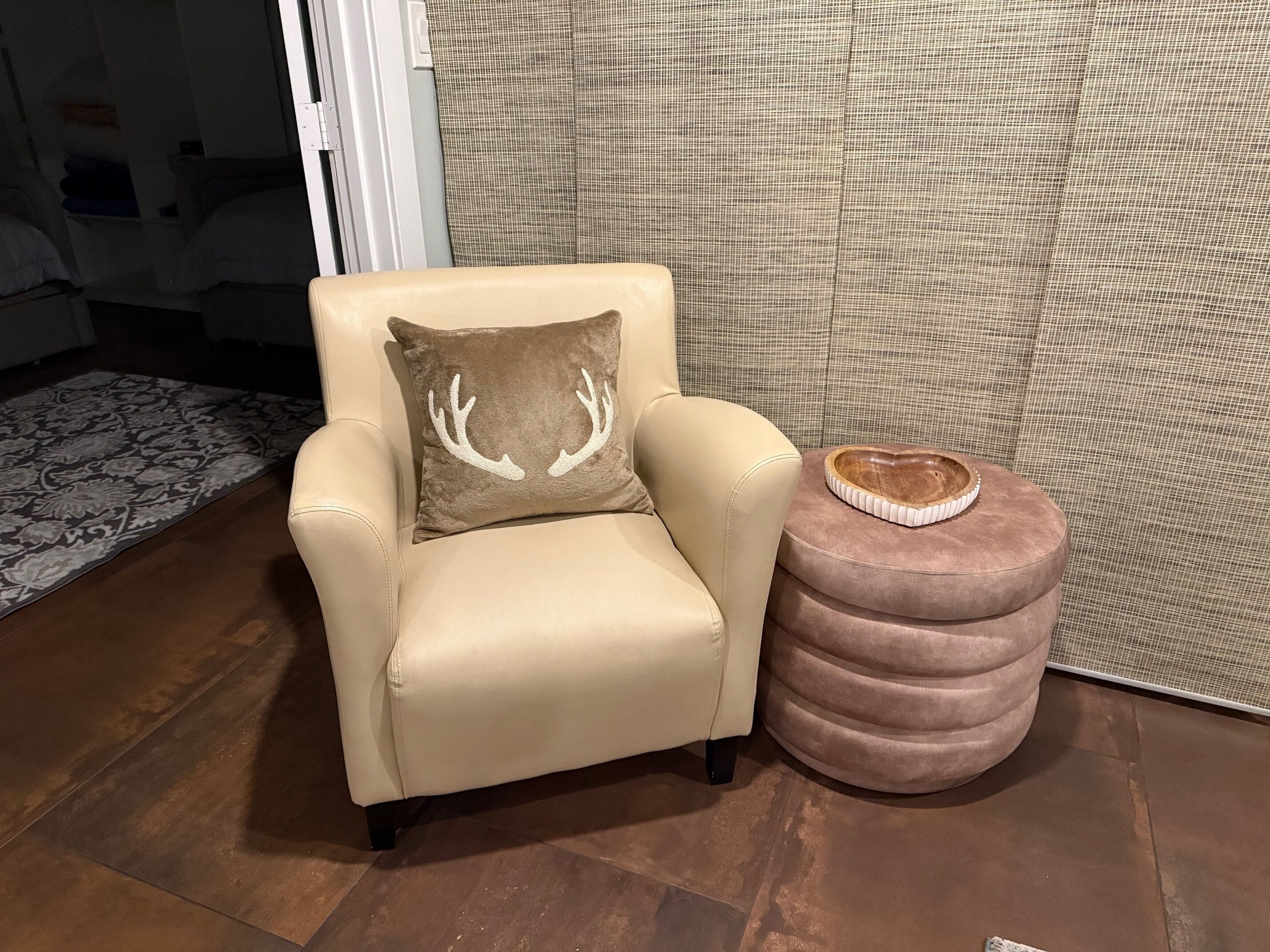 Un sillón beige con un cojín de astas de ciervo, un reposapiés redondo con una bandeja de madera, y una alfombra estampada sobre un piso de madera.