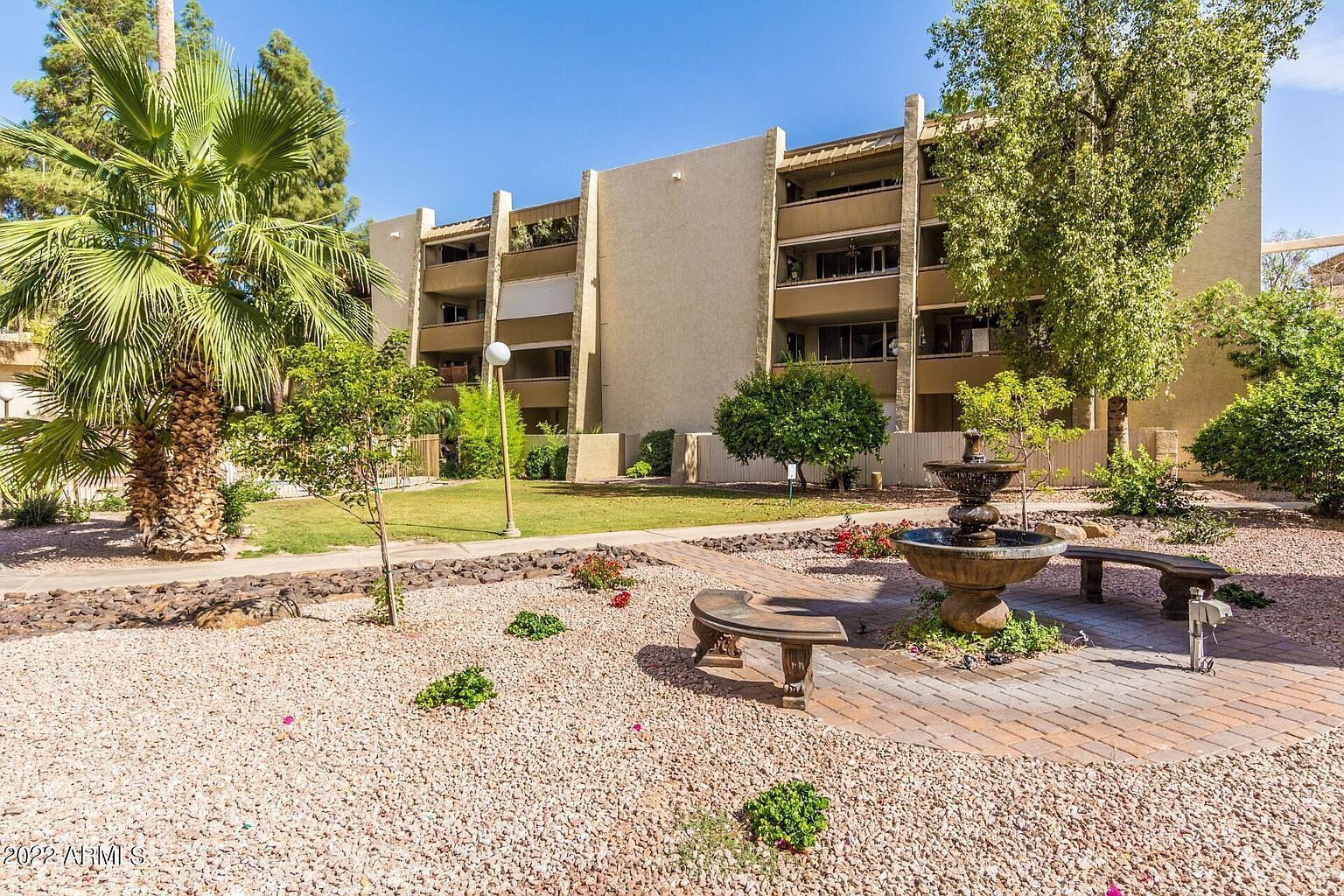 A multi-story building with balconies, surrounded by a landscaped garden with palm trees, a fountain, and a paved walkway.