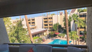 Un balcó amb cadires amb coixins blancs i almoades vermelles i blaves, amb vistes a una piscina i una zona enjardinada amb palmeres i un edifici al fons.