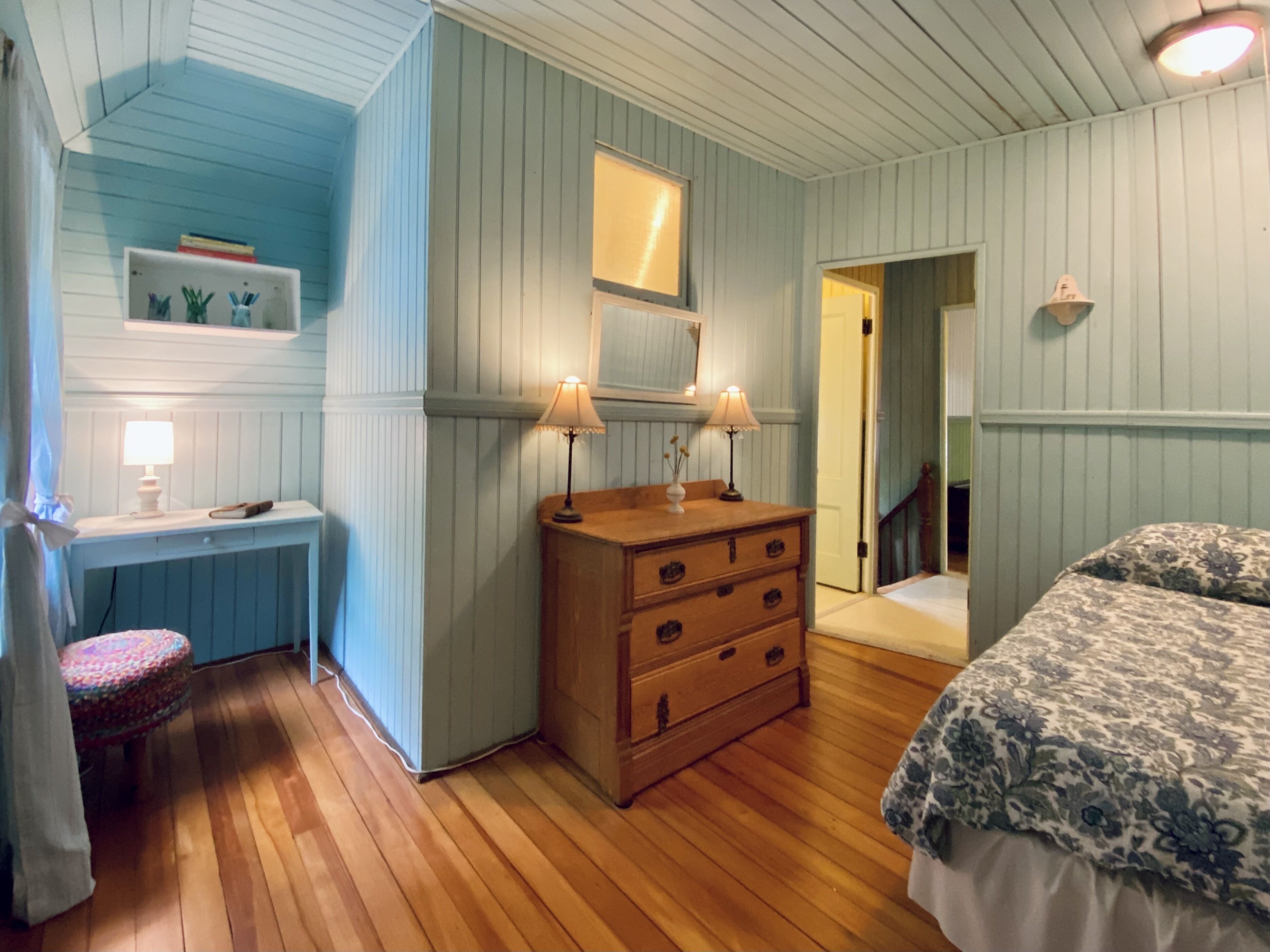 Une chambre avec une commode en bois, un lit recouvert d’une couvre-lit à motifs floraux, une coiffeuse avec un miroir et une petite table avec une lampe.