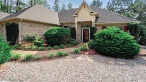 A two-story house with a stone facade, a front porch, and a well-maintained garden with various shrubs and a gravel driveway. - Quiet, Beautiful Home Nestled in the woods (Hot Springs Village)