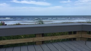 Une terrasse en bois donnant sur une plage, avec des vagues et un ciel dégagé.