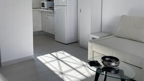 A modern, minimalist kitchen with white cabinetry and appliances, a glass coffee table with a bowl, and a white sofa in a bright room with natural light.