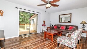 Un salon doté d’un ventilateur de plafond, d’un canapé en cuir marron, d’un fauteuil à motifs, d’une table basse en bois et d’une porte-fenêtre coulissante donnant sur une terrasse extérieure.
