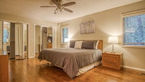 Ein Schlafzimmer mit Holzboden, einem Bett mit grauer Bettdecke, einem Deckenventilator, einem Nachttisch mit Lampe und einem Fenster mit Jalousien.