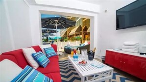 Una sala de estar moderna con un sofá rojo, una mesa de centro blanca y un mueble de almacenamiento rojo. Hay un televisor montado en la pared y vistas a un área exterior con sombrillas.