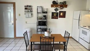 A dining area with a wooden table and four chairs, a kitchen with a refrigerator and stove, and a shelf with kitchenware and decorations on the wall.