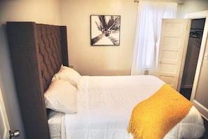Una cama bien hecha con ropa de cama blanca y una manta amarilla, un cabecero oscuro, un cuadro enmarcado en la pared y una puerta de armario entreabierta.