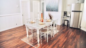 Uma área de refeições com uma mesa branca posta para quatro pessoas, rodeada por cadeiras brancas. Um quadro grande está na parede acima da mesa.