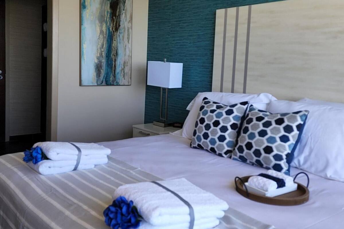 Quarto de hotel com uma cama grande, cabeceira, um quadro na parede, mesa de cabeceira com abajur e toalhas dobradas na cama.
