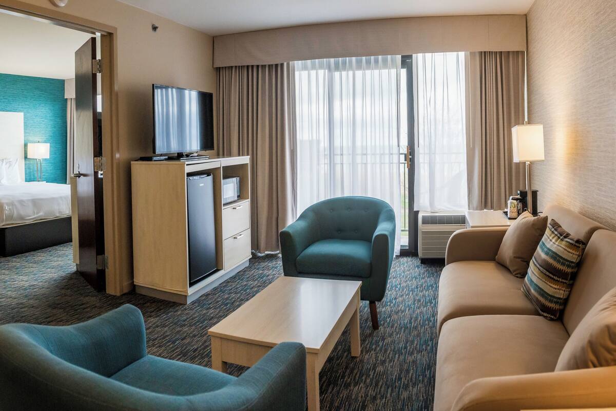 Quarto de hotel com cama, escrivaninha com computador, televisão, sofá e poltrona.