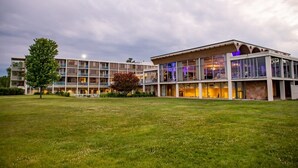 Un hotel moderno con varios pisos, amplias ventanas de vidrio y un área exterior espaciosa con césped y árboles.