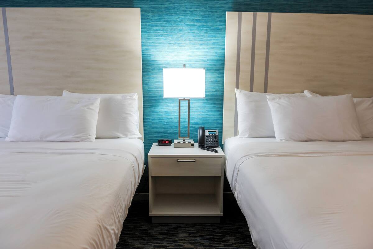 Quarto de hotel com duas camas, mesa de cabeceira com abajur, telefone e relógio digital.
