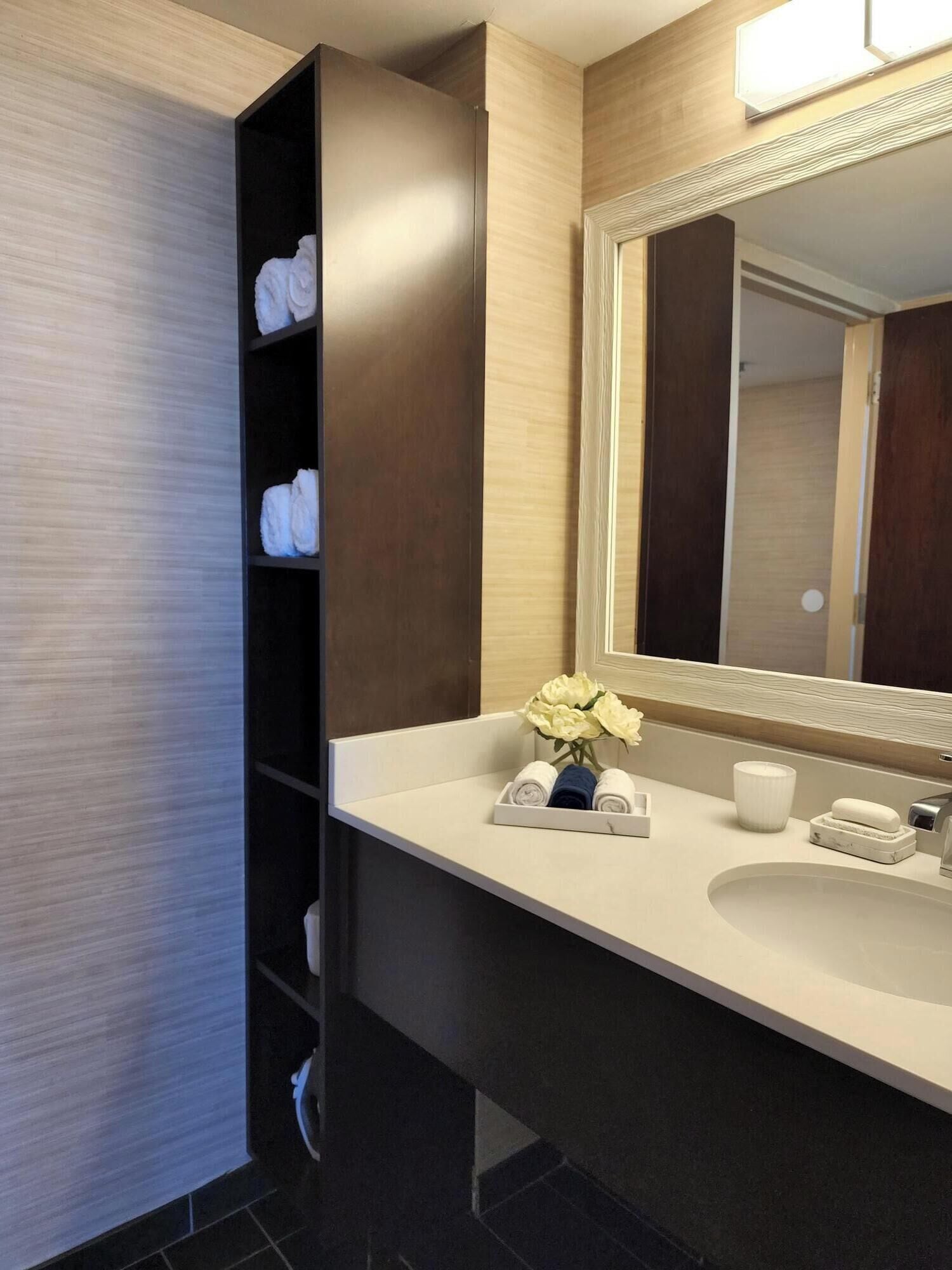 A modern bathroom with a dark wood vanity, a large mirror, and neatly folded towels on shelves.