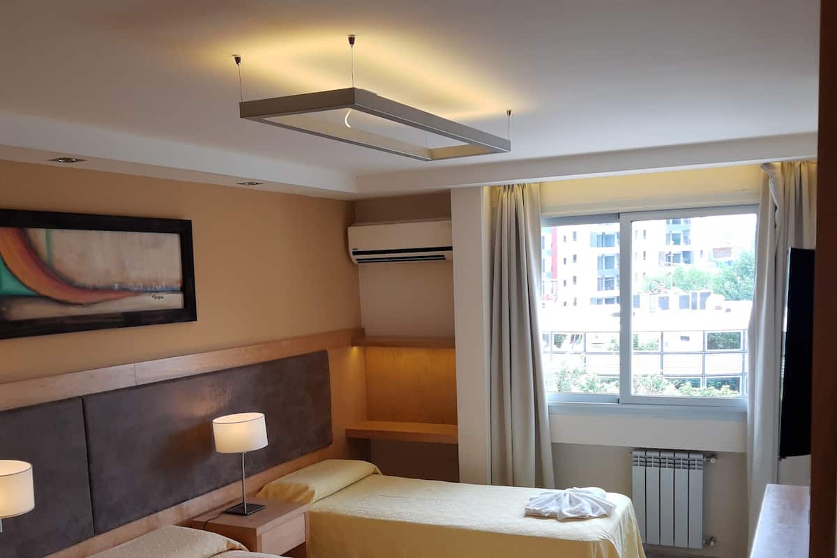 Une chambre d’hôtel avec deux lits, un tableau au mur, un appareil de climatisation, une fenêtre avec des rideaux et une vue sur les bâtiments environnants.