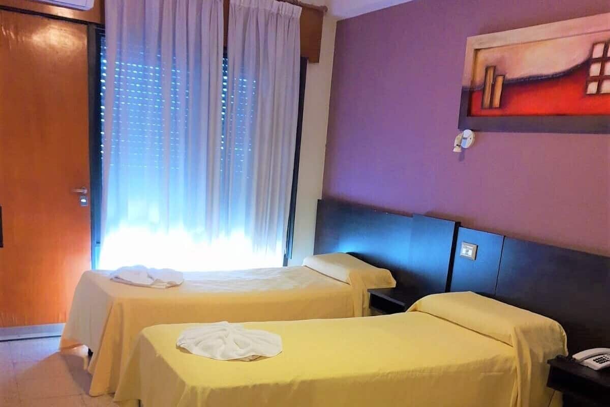 Une chambre d’hôtel avec deux lits, chacun recouvert d’une couverture jaune et agrémenté d’une serviette blanche. Il y a une fenêtre avec des rideaux, un tableau au mur et un téléphone sur la table de chevet.