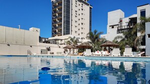 En swimmingpool med liggestole og parasoller, omgivet af bygninger og palmer.