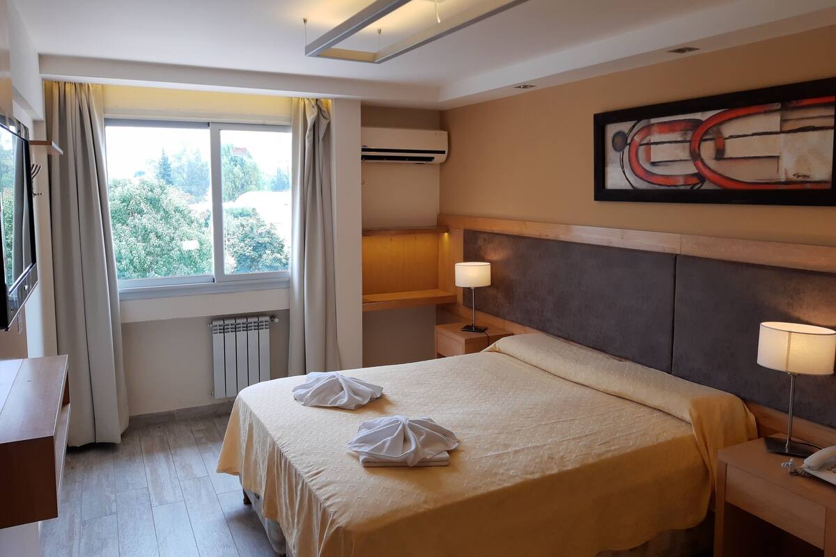 Une chambre d’hôtel avec un lit, deux lampes de chevet, une fenêtre avec des rideaux, un radiateur, un climatiseur mural et un tableau abstrait encadré.