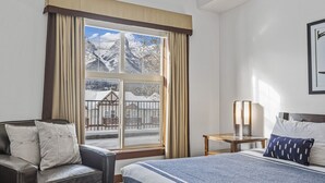 Habitación de hotel con cama, un sillón de cuero, una mesita de noche con lámpara y ventana con vista a un paisaje nevado.