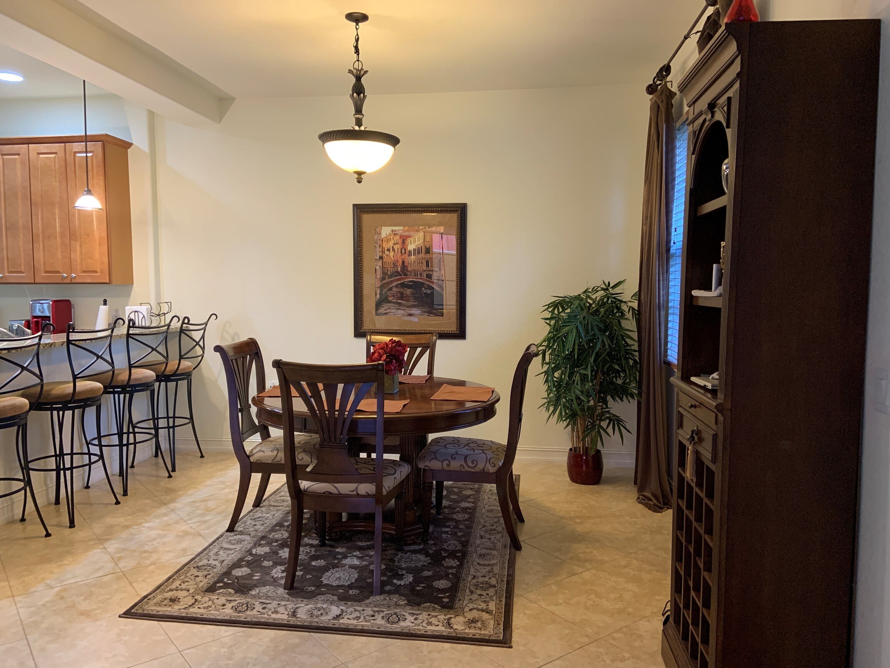 A dining area with a round table, chairs, a patterned rug, a hanging light fixture, and a framed picture on the wall.