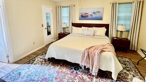A hotel room with a large bed, two bedside tables, a ceiling fan, a window with curtains, and a painting on the wall. - Golf Cart to Beach, Pet Friendly, Fenced Yard, Amelia Island Quiet Home Escape (Fernandina Beach)