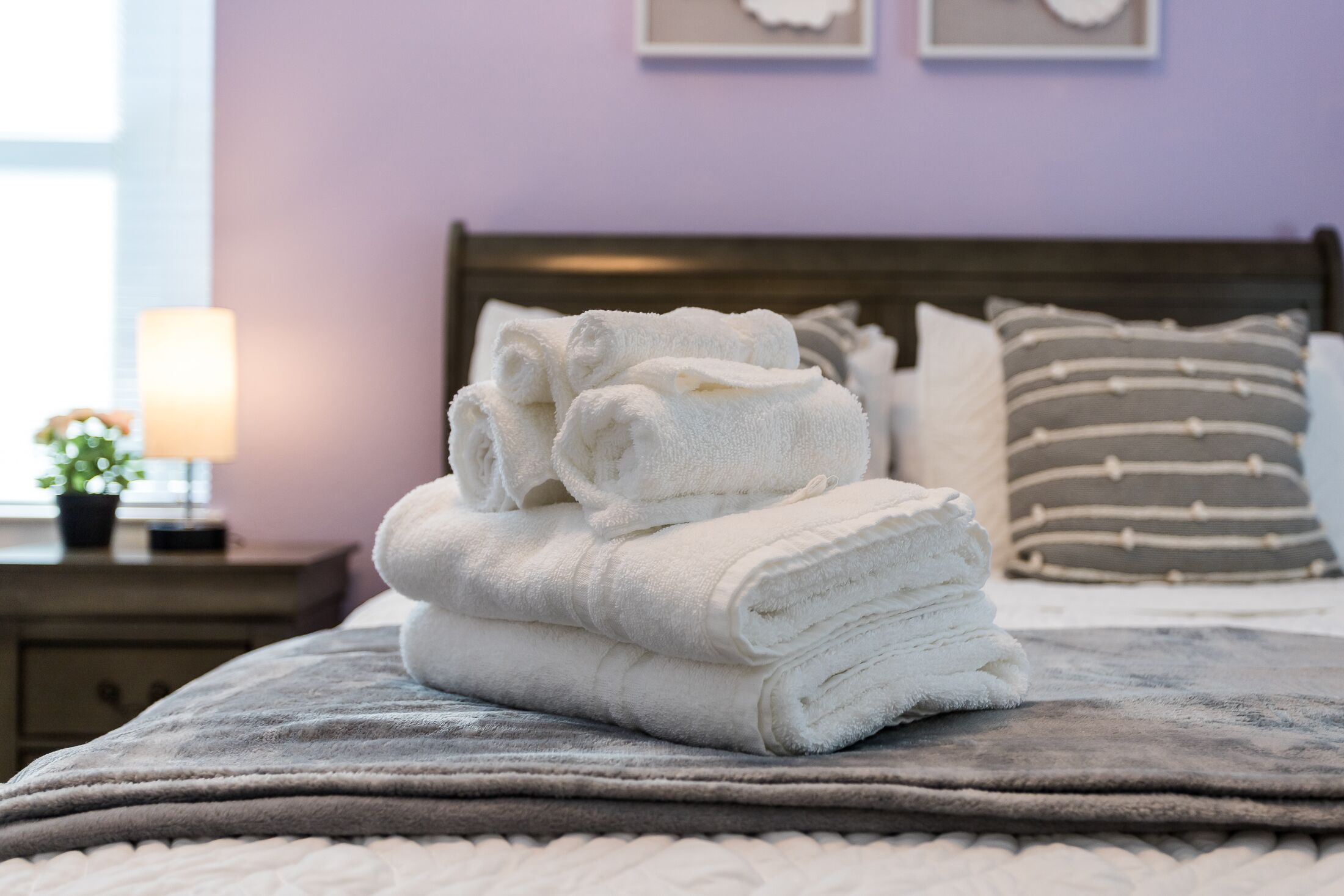 A neatly made bed with a stack of white towels on a gray blanket, a bedside lamp, and a nightstand with a small plant.