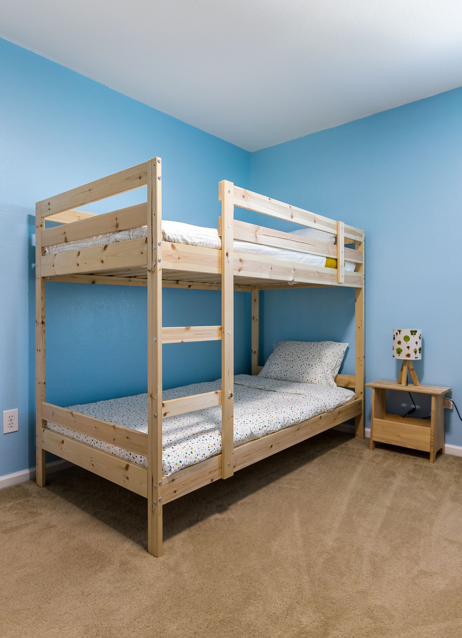 A bunk bed with a wooden frame, a small bedside table, and a wall socket.