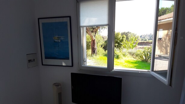 A room with a bed, a TV on a stand, a window with a view of greenery, and a framed picture on the wall. - Maison Petit Jardin au Calme Parking (Afa)
