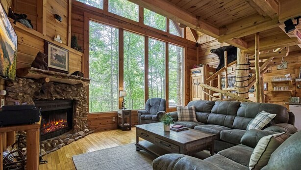 L’intérieur chaleureux d’un chalet avec une cheminée en pierre, un canapé en cuir et de grandes fenêtres donnant sur la forêt.
