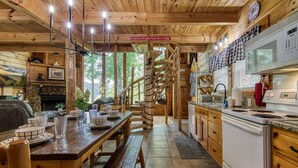 L’intérieur d’un chalet rustique comprend une table à manger en bois pour quatre personnes, une cuisine équipée d’une cuisinière et d’un évier, ainsi qu’un escalier en colimaçon menant à un étage supérieur.