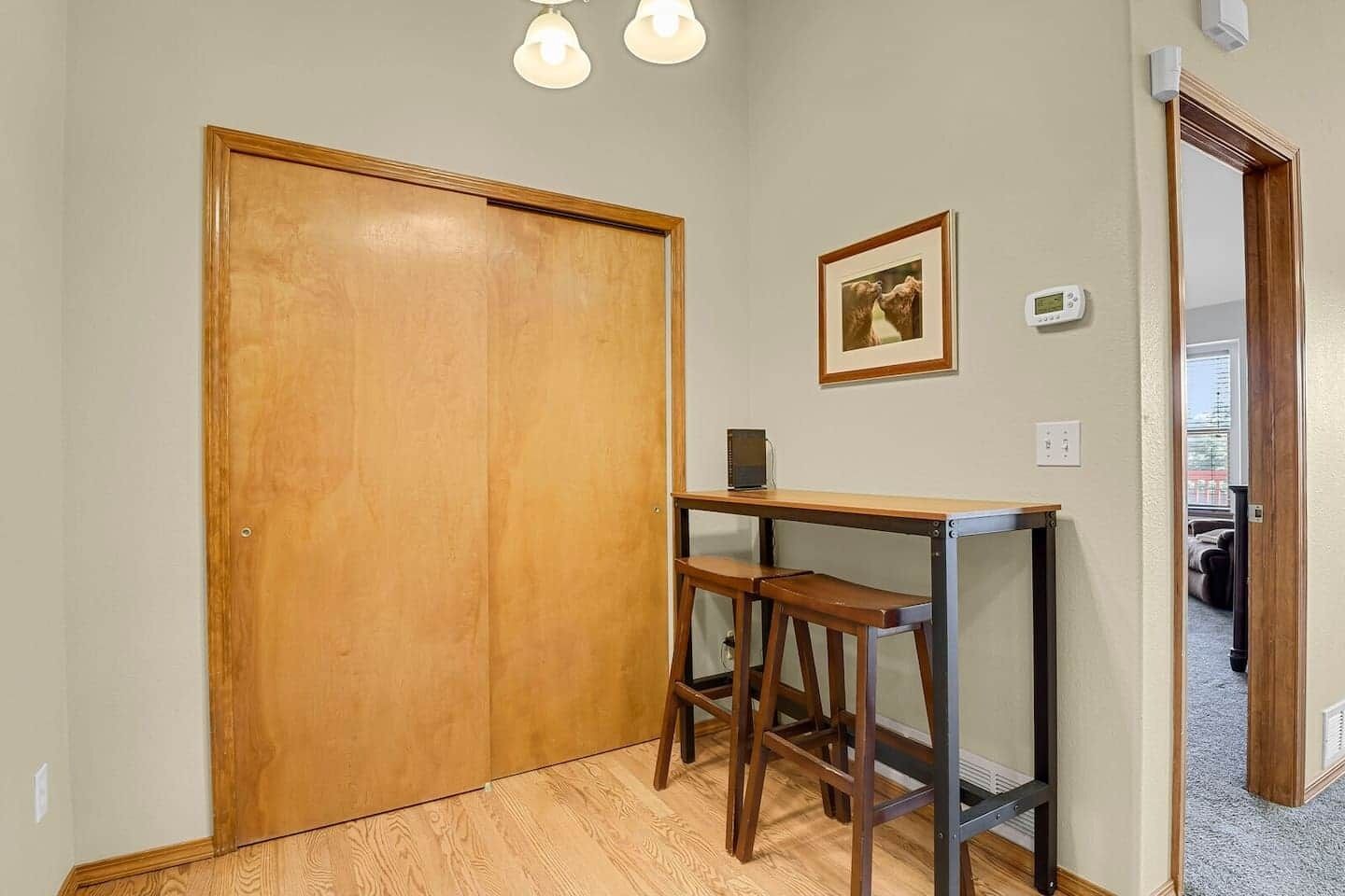 A room with a wooden door, a small table with two stools, a framed picture on the wall, and a thermostat.
