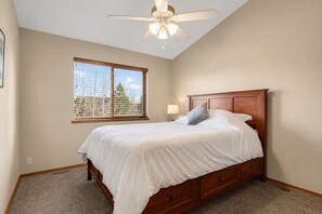 Une chambre à coucher comprenant un lit, un ventilateur de plafond, une fenêtre avec des stores et une table de chevet avec une lampe.