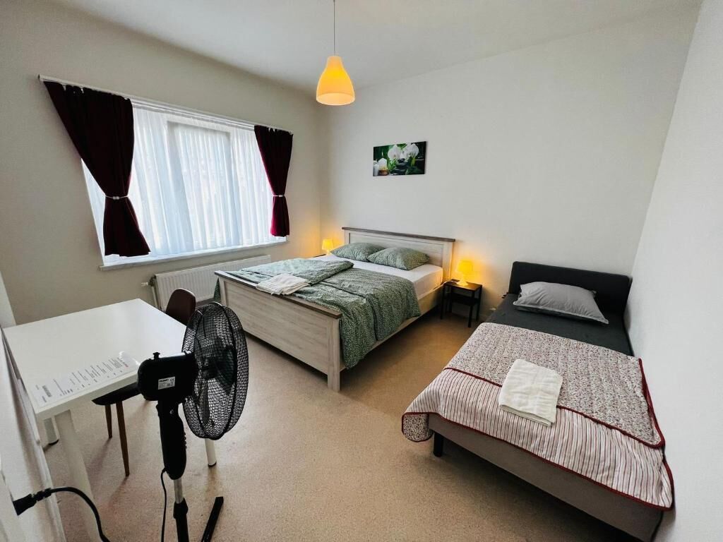 Un dormitorio compacto con una cama individual, una cama más pequeña, un escritorio y un ventilador de pie.