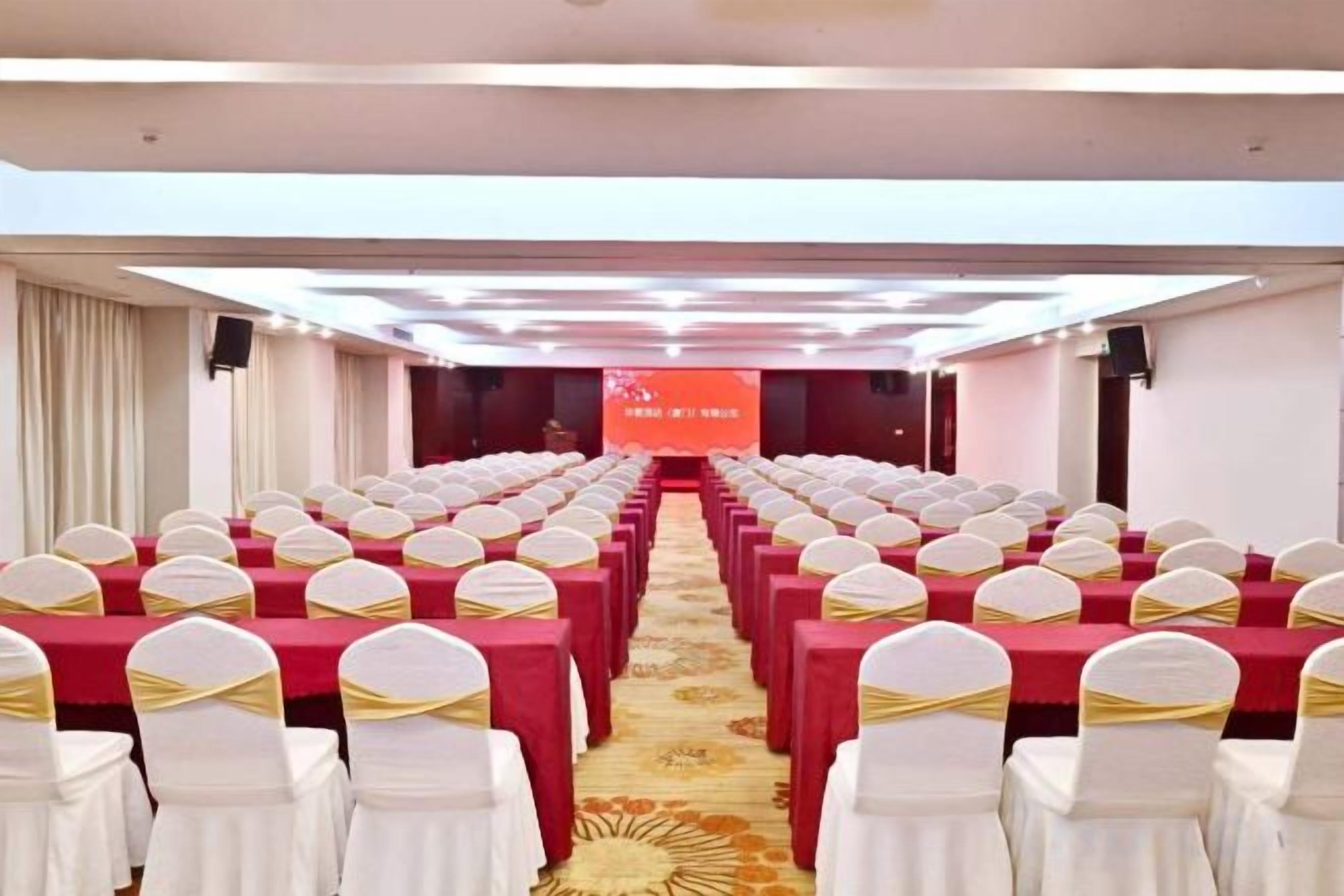 A conference room with rows of tables and chairs, a stage with a screen, and a carpeted floor.