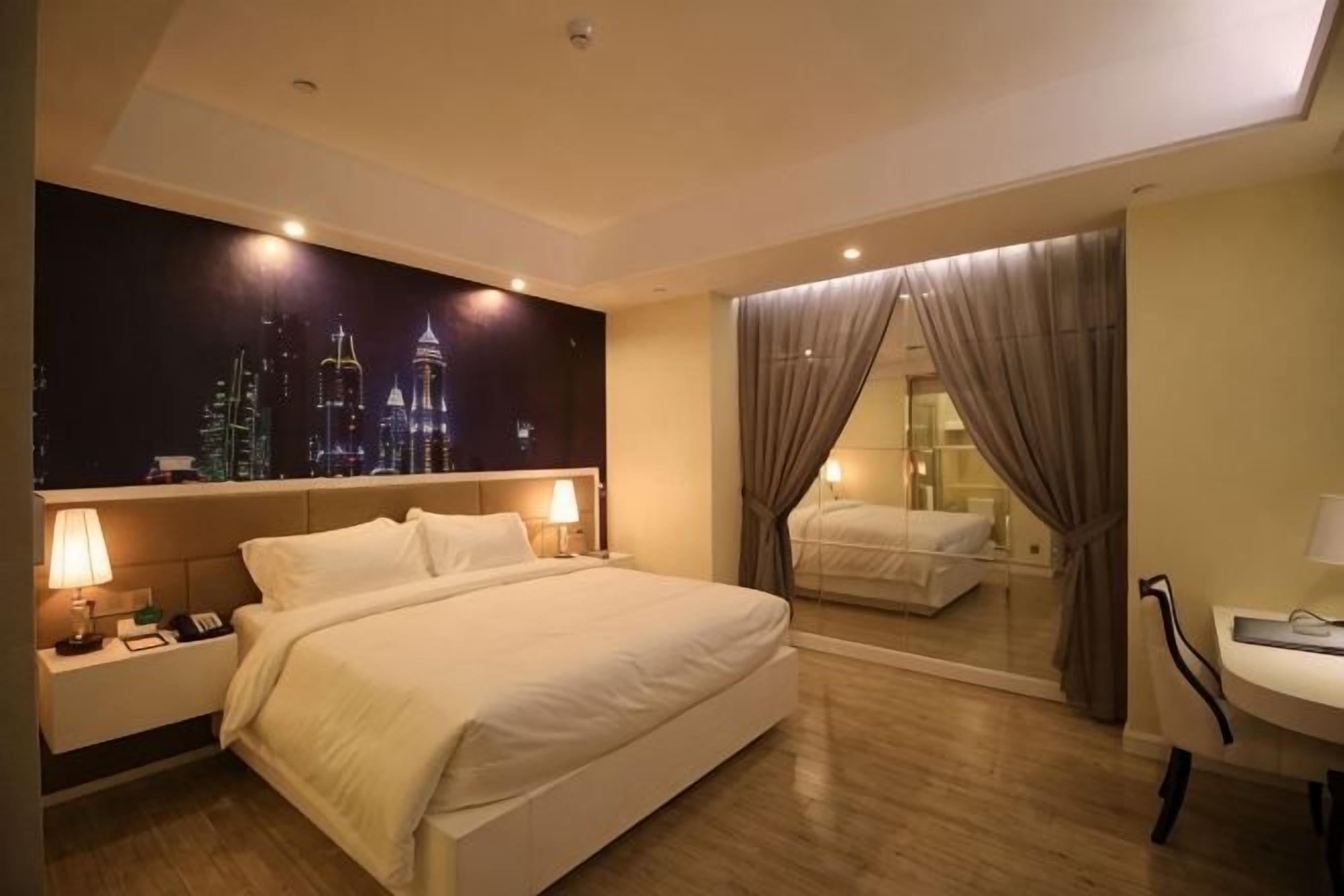 A hotel room with a large bed, bedside tables, a nightstand, a mirror, and a view of the cityscape.