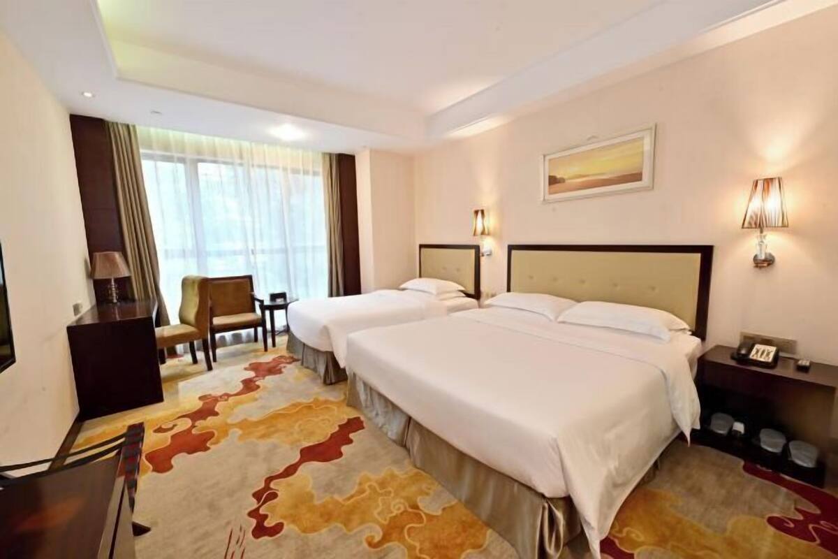 A hotel room with two beds, a desk, a chair, a lamp, and a painting on the wall.