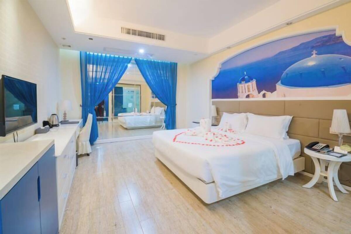 A hotel room with a bed, a TV, a desk, and a painting of a building on the wall.