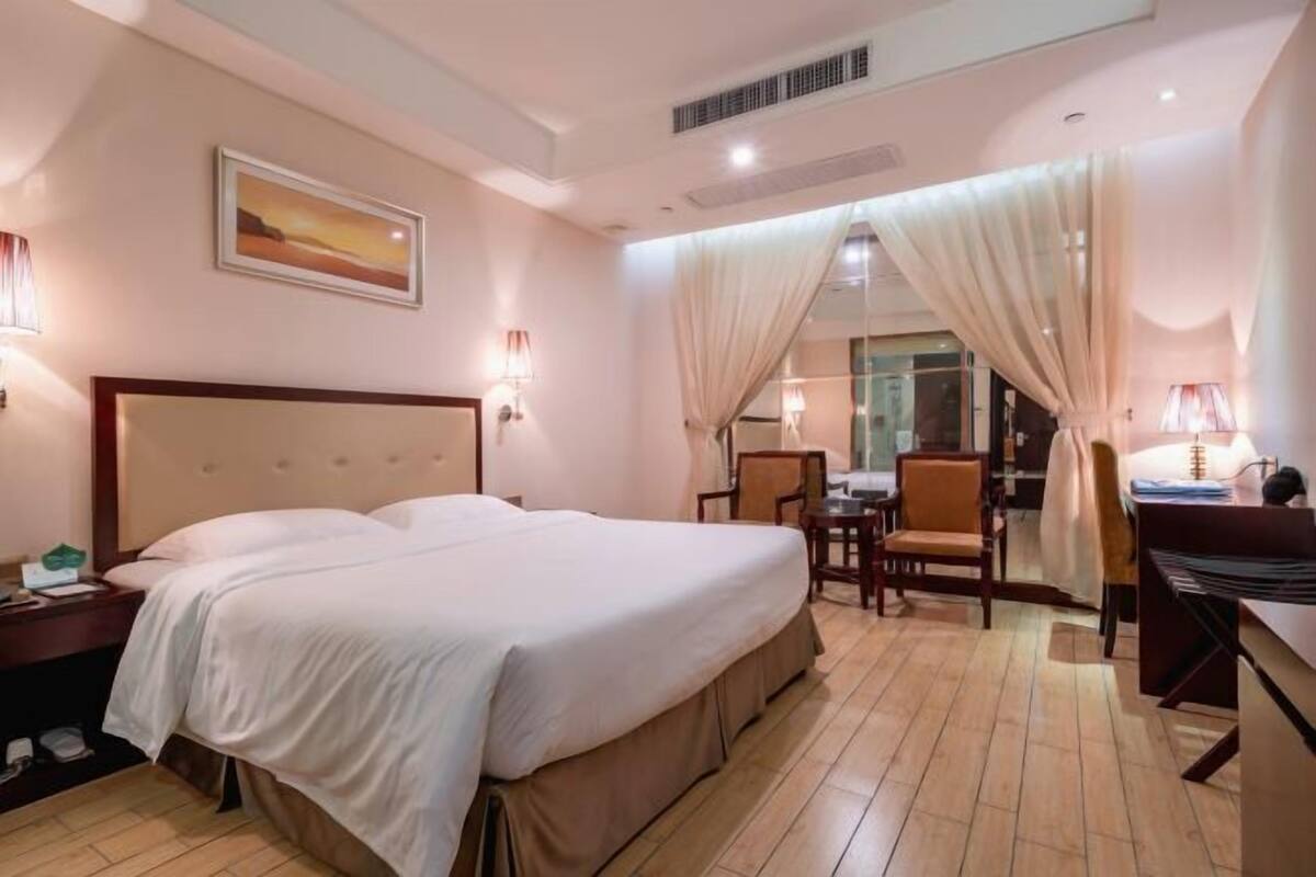 A hotel room with a large bed, a desk, and a chair. There is a painting on the wall and a window with curtains.