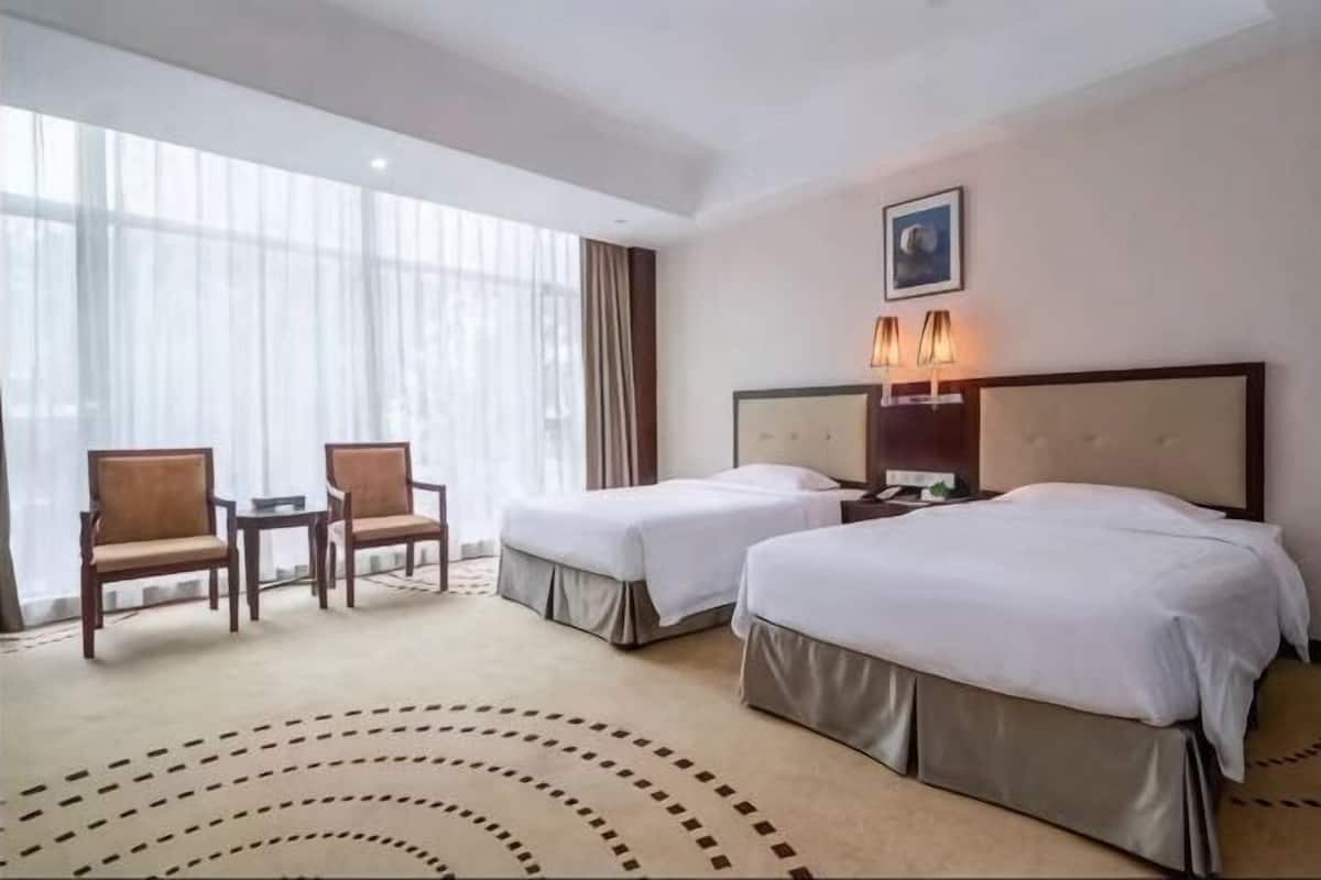 A hotel room with two beds, a desk, chairs, and large windows.