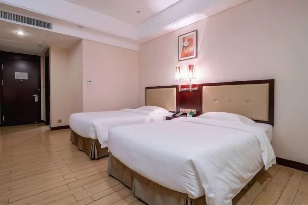 A hotel room with two beds, a wooden floor, a door, and a wall-mounted light fixture.