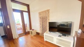 Sala de estar con una televisión de pantalla plana sobre un mueble blanco, piso de madera, puerta corrediza de vidrio que da al exterior y una pequeña mesita con un cuenco.
