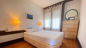 Una cama bien hecha en una habitación con suelo de madera, un aparador y dos lámparas fijadas a la pared.