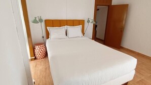 Un llit ben fet a una habitació amb terra de fusta, una porta tancada i una taula rodona petita amb un tamboret de disseny geomètric.
