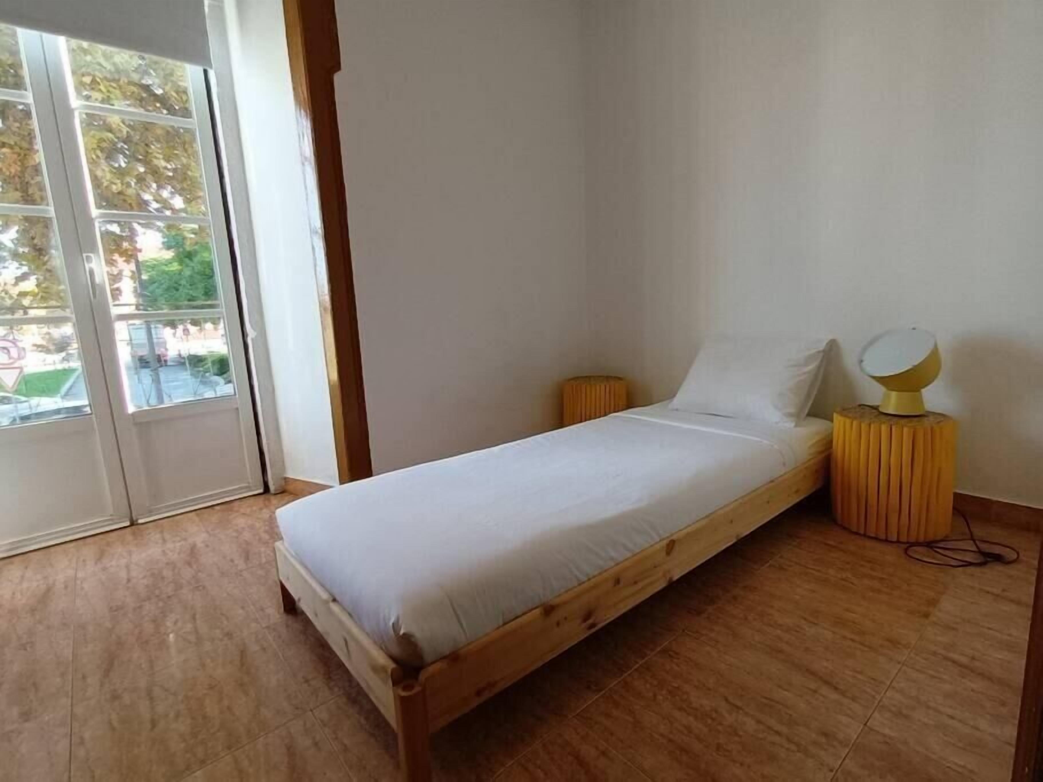 A bedroom with a wooden bed, a small bedside table with a lamp, and a sliding glass door leading to an outdoor area.
